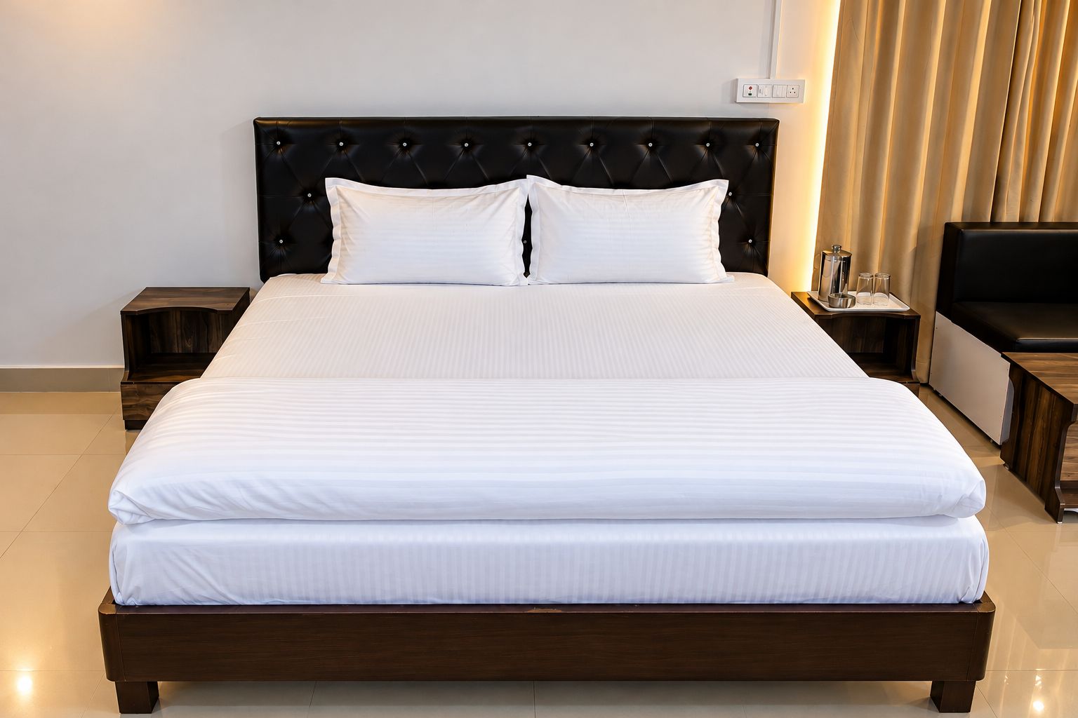 Una cama grande con cabecera oscura y ropa de cama blanca, flanqueada por mesitas de noche de madera con un juego de vasos y una pequeña mesa.