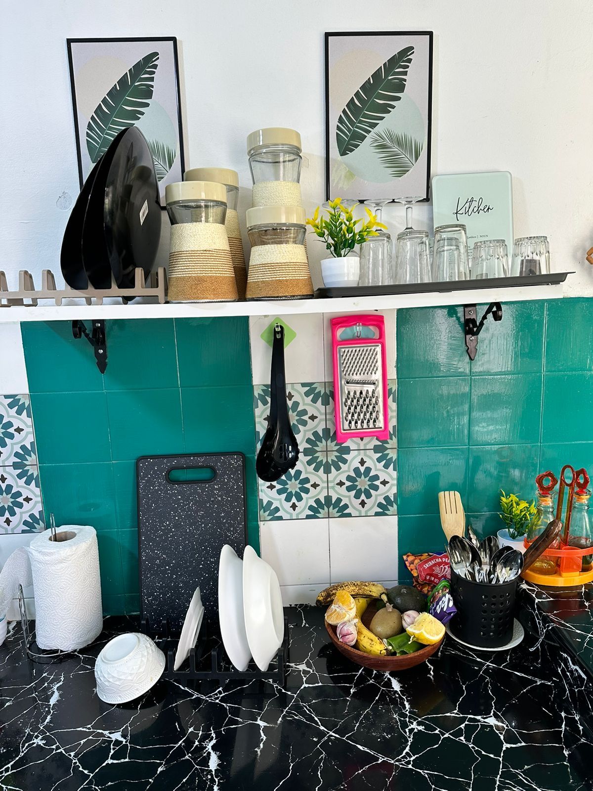 A kitchen counter with various utensils, a cutting board, a bowl of fruit, and a hanging kitchen tool.