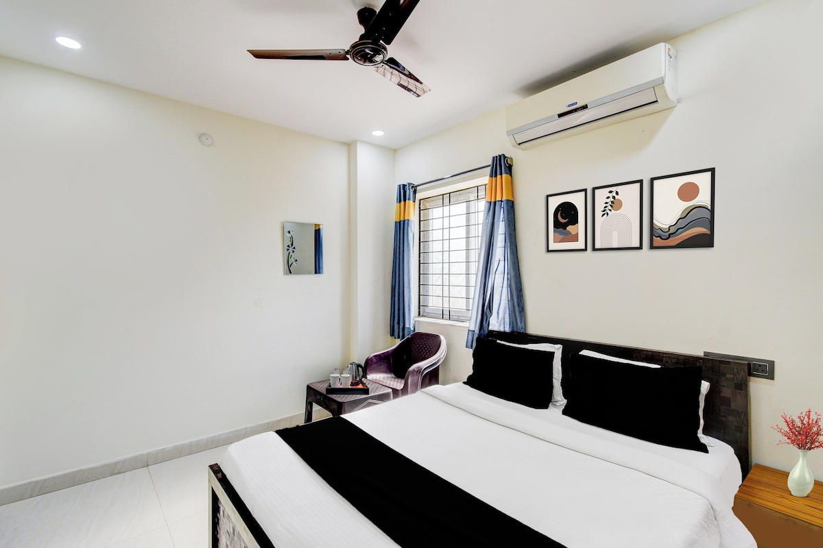 Una habitación de hotel con cama, una silla, una mesa con una bandeja de objetos, un equipo de aire acondicionado, una ventana con cortinas y una obra de arte en la pared.