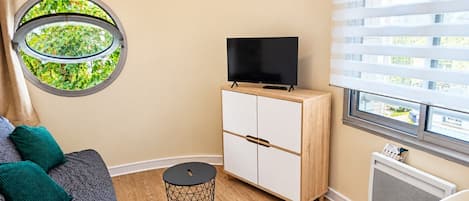 Una habitación con un ventanal redondo, un televisor sobre un mueble de madera, un sofá con cojines, una mesita redonda pequeña y un radiador.