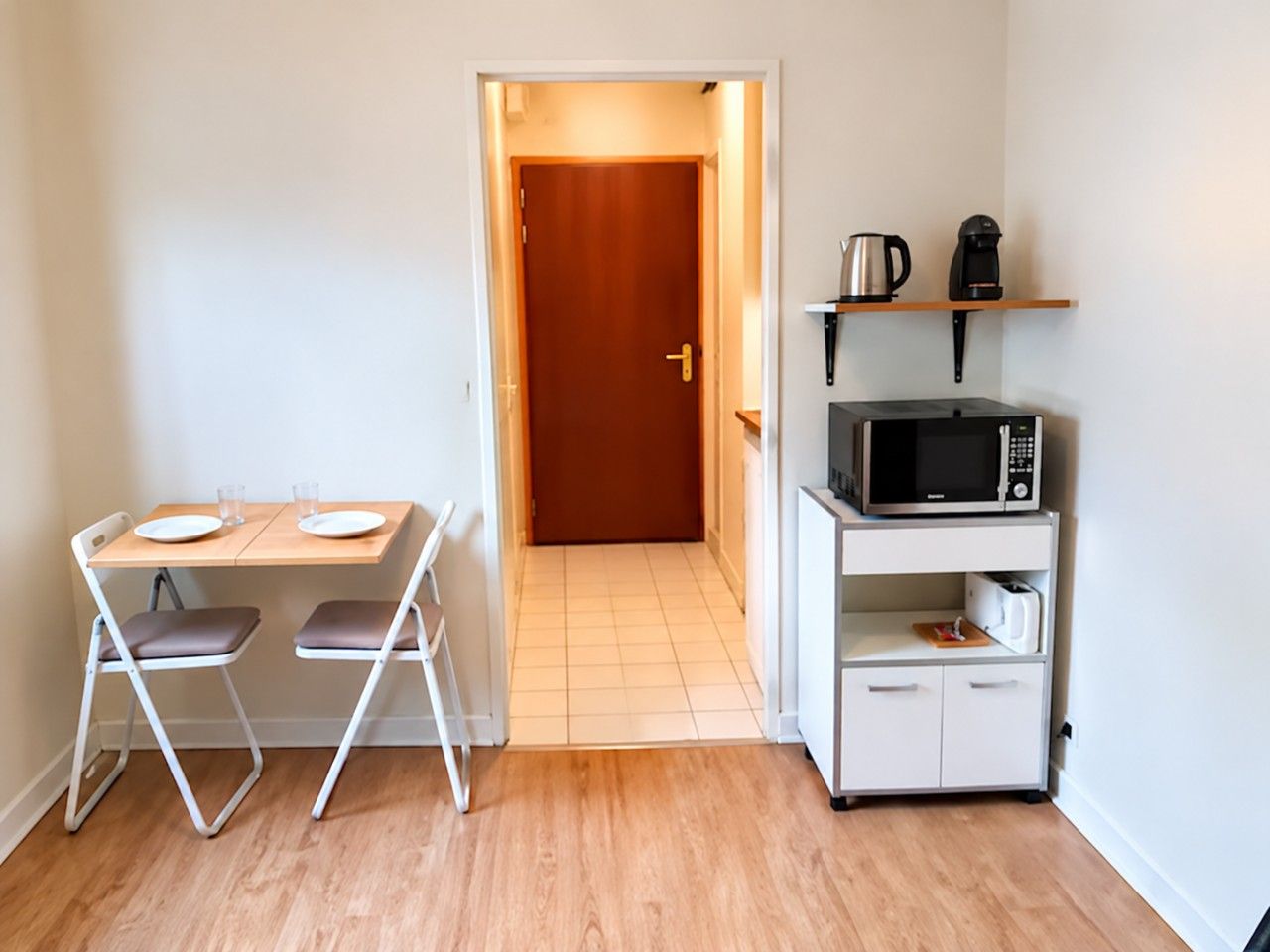 A compact kitchen with a microwave, toaster, and coffee maker on a shelf, a small table with two chairs, and a door leading to another room.