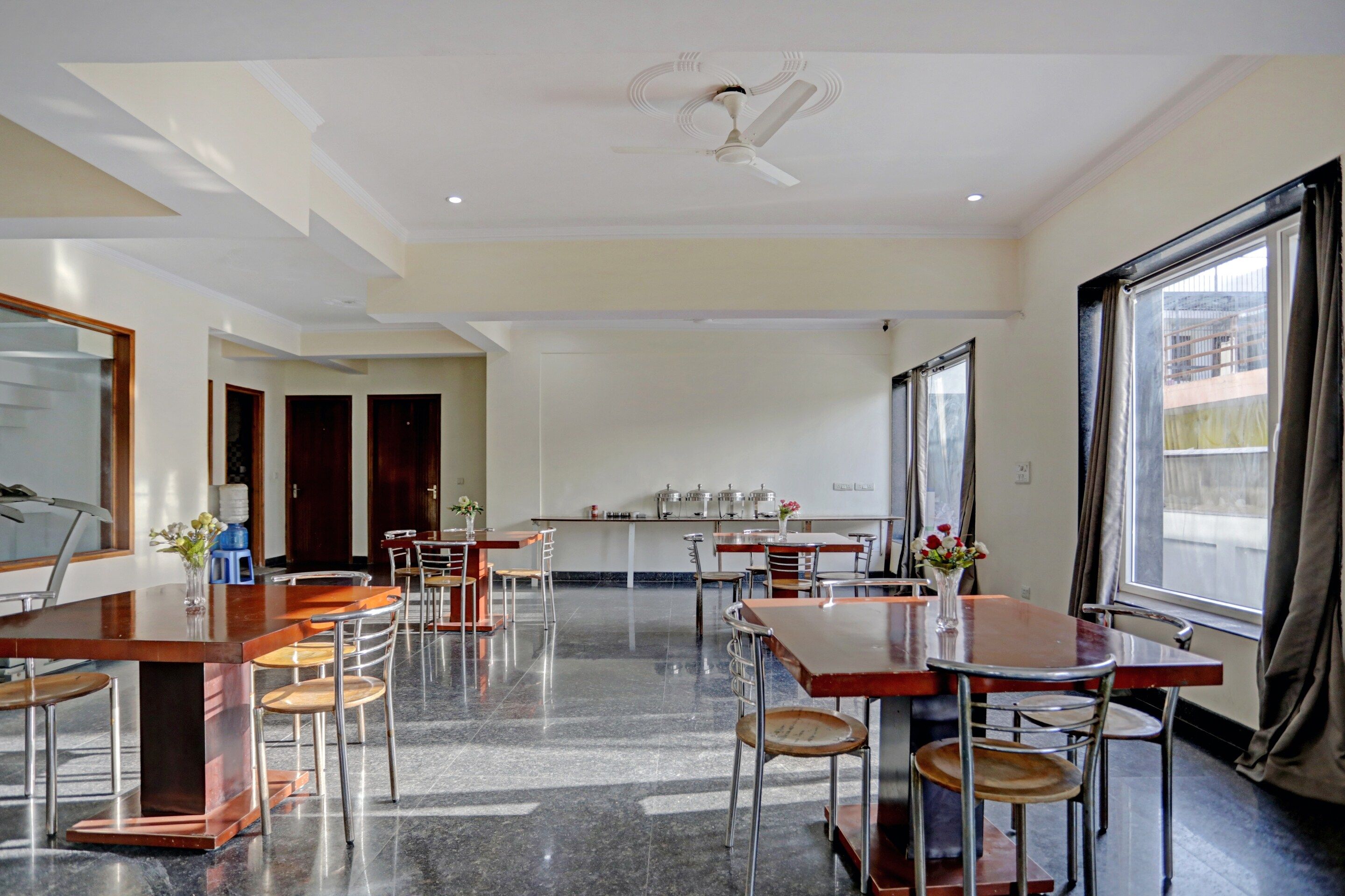 A spacious dining area with multiple tables and chairs, a ceiling fan, and large windows with curtains.