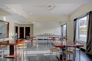 A spacious dining area with multiple tables and chairs, a ceiling fan, and large windows with curtains. - Townhouse Merriment Near ISKCON Temple (Noida)