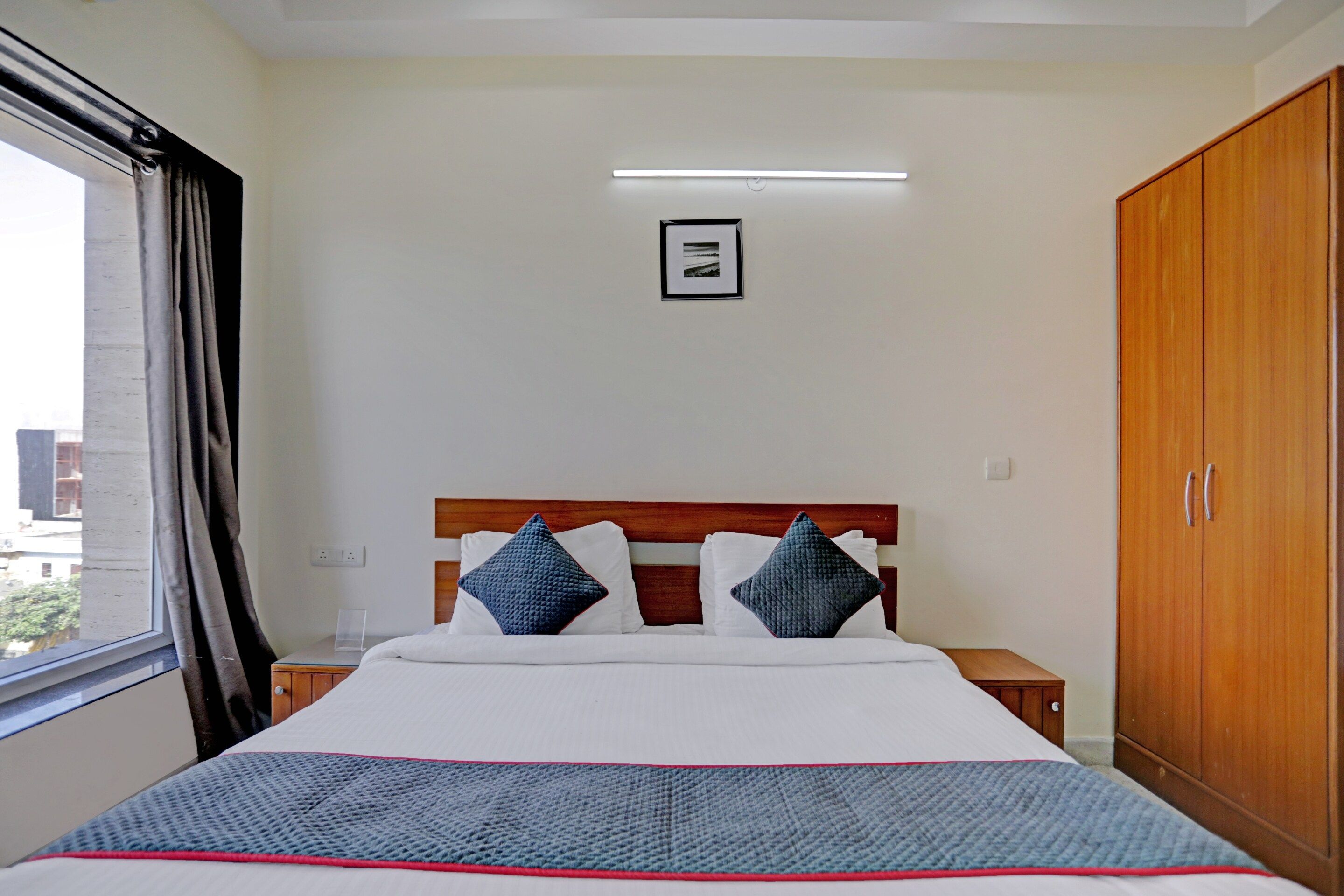 Habitación de hotel con cama, mesita de noche, armario, ventana con cortinas y lámpara fijada a la pared.