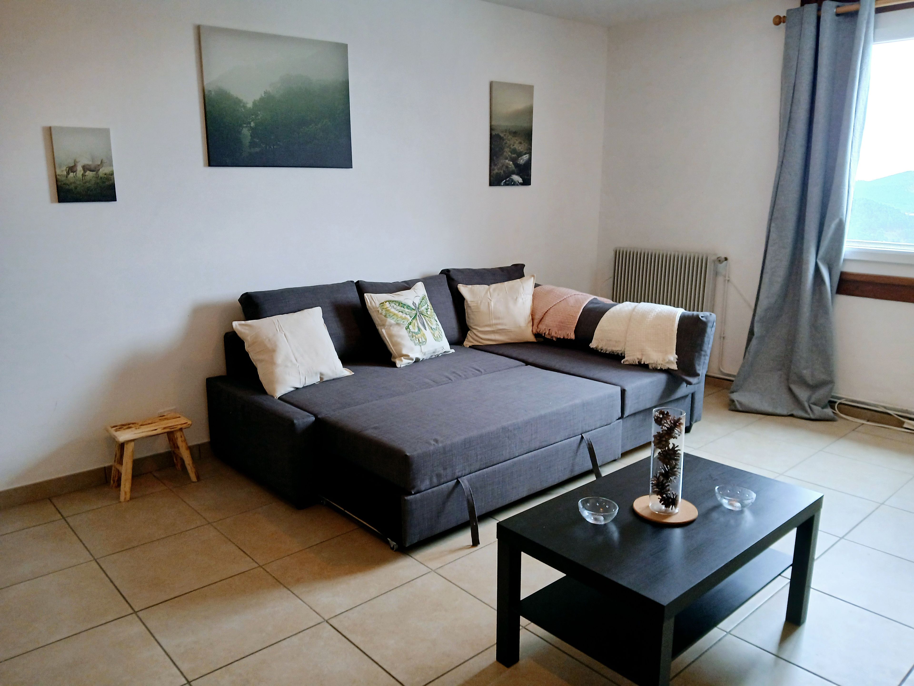 A modern living room with a sofa, a small wooden stool, a glass table with decorative items, and a window with curtains.