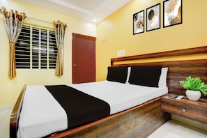 Habitació d'hotel amb un llit, una tauleta de nit, una planta en test i una finestra amb cortines.