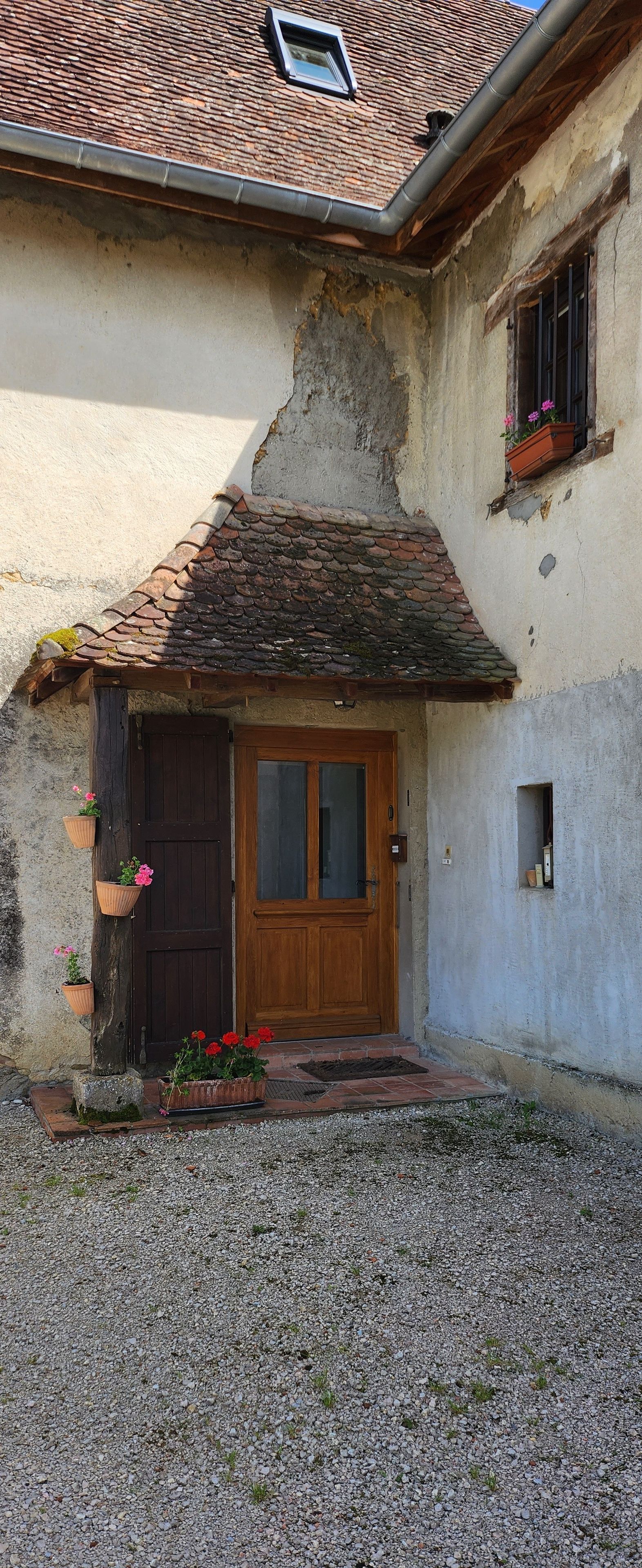 A rustic building with a wooden door, a small window with flowers, and a gravel driveway.
