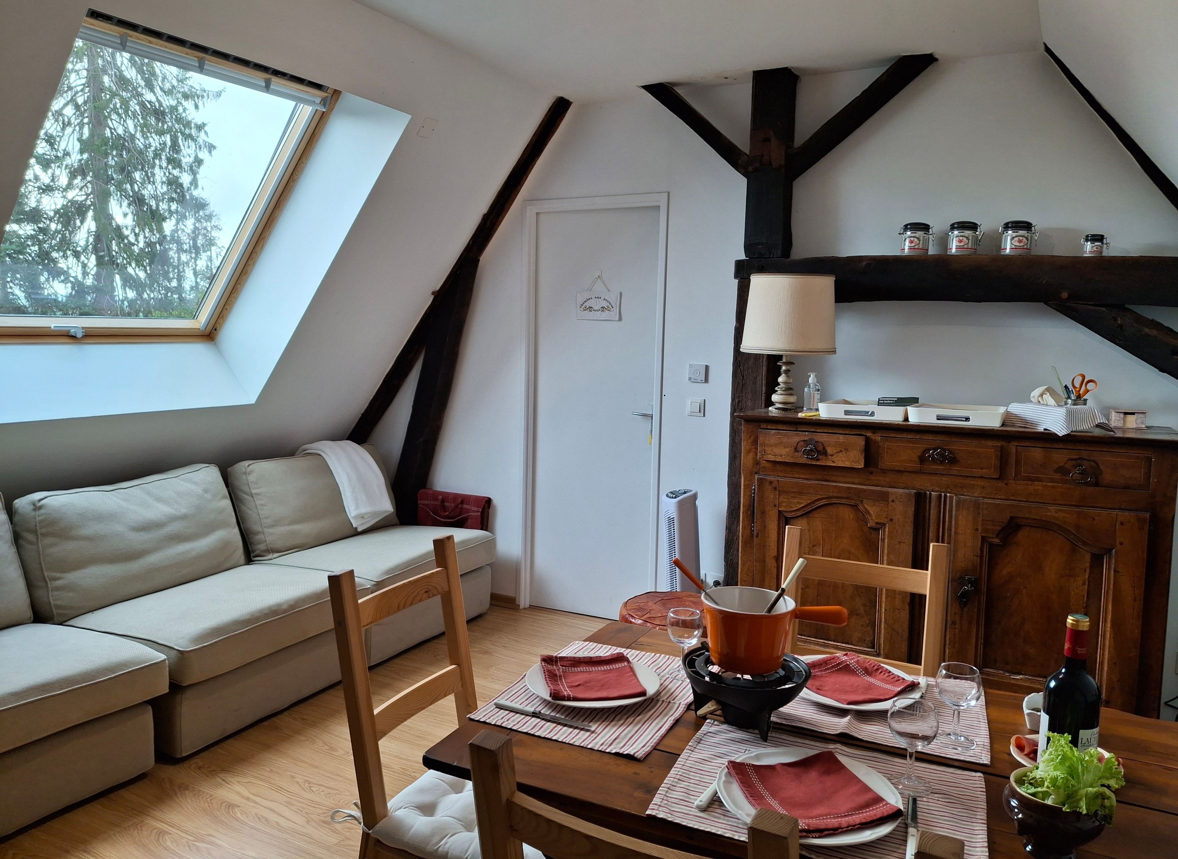 A cozy loft bedroom with a sloped ceiling, a skylight, a wooden dining table set for two, a sofa, and a cabinet with kitchenware.