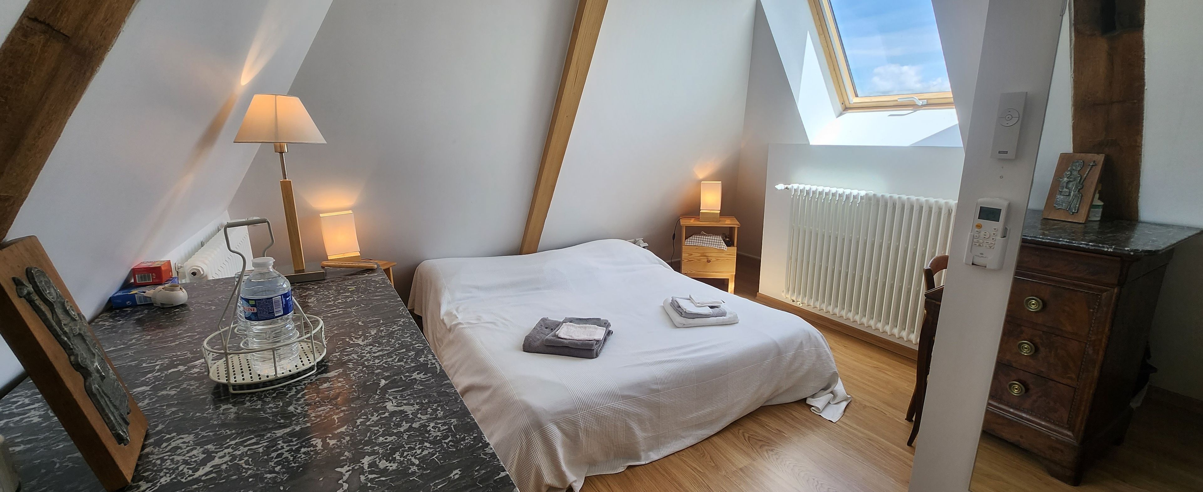 A bedroom with a skylight, a bed with white bedding, a nightstand with a lamp, a wooden dresser, and a water dispenser on the counter.