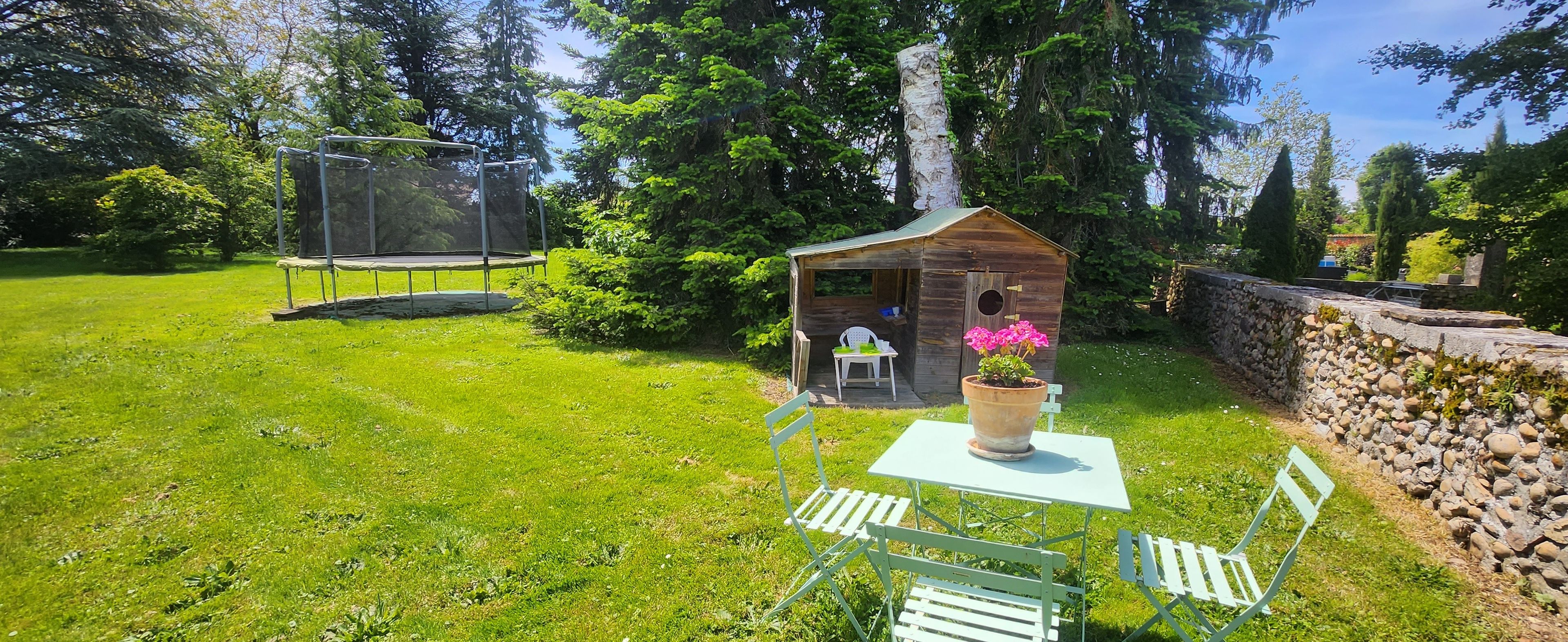 A garden with a trampoline, a wooden structure with a window, and a table with chairs.