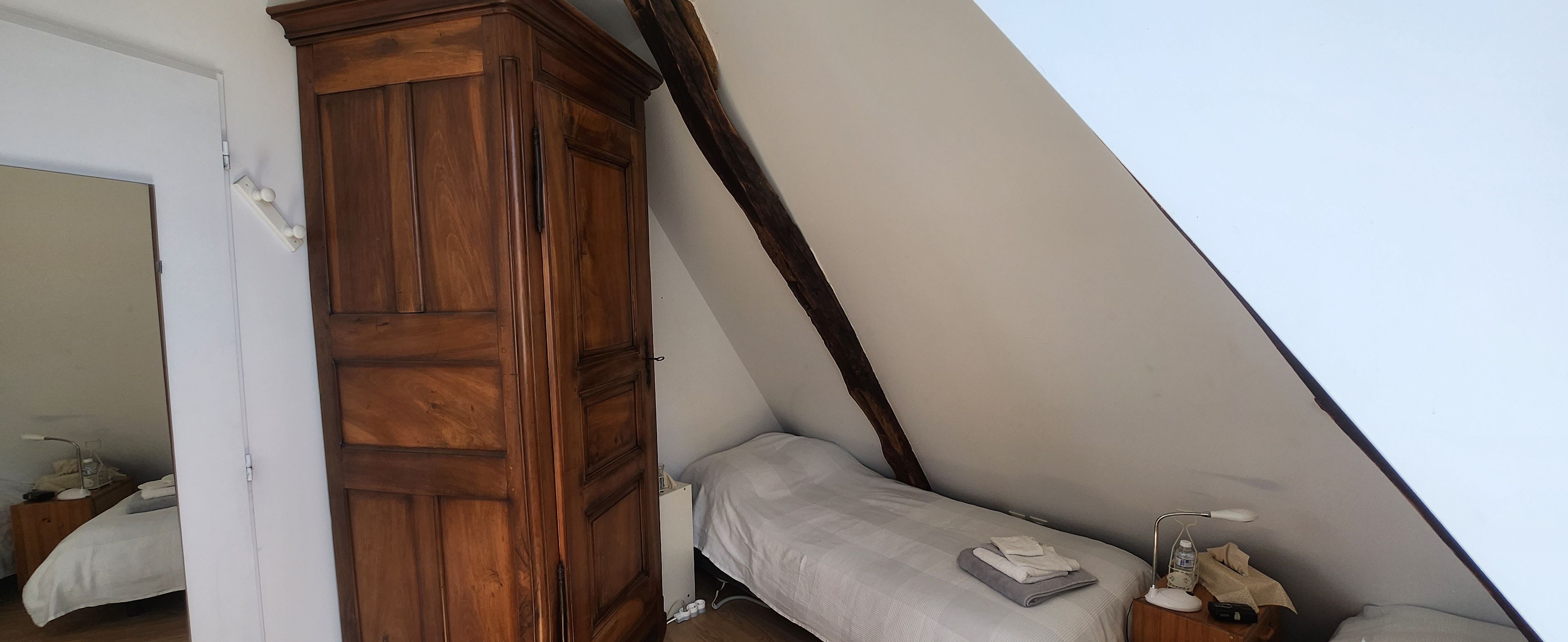 A bedroom with a wooden wardrobe, a single bed, a bedside table with a lamp, and a window.