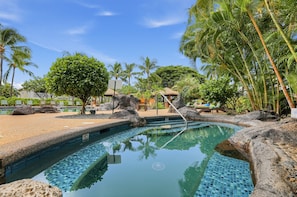 Ein klares Schwimmbecken mit einer gefliesten Kante, umgeben von Felsen und Palmen, mit Liegebereich und einer strohgedeckten Struktur im Hintergrund.