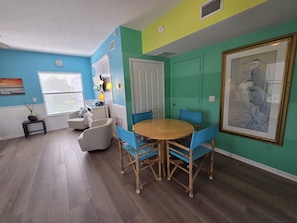 A room with a round wooden table, blue chairs, a white door, and a framed bird painting on the wall. - Siesta Sunset Royale #6 1 Bedroom Apts (Siesta Key)