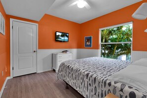 Een slaapkamer met een oranje muur, een witte deur, een bed met een bedovertrek met een patroon, een dressoir, een televisie aan de muur en een raam met uitzicht op groen.