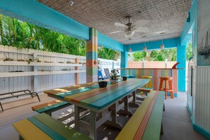 An outdoor dining area with colorful benches and a table, surrounded by a white fence and lush greenery. - Siesta Sunset Royale #1 1 Bedroom Apts (Siesta Key)