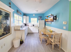 A kitchen with white cabinets, a dining table with chairs, a refrigerator, and a television mounted on the wall. - Siesta Sunset Royale #1 1 Bedroom Apts (Siesta Key)