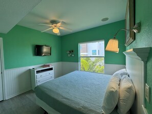 Un dormitorio con cama, ventilador de techo, ventana con vistas, televisor en la pared y cómoda.