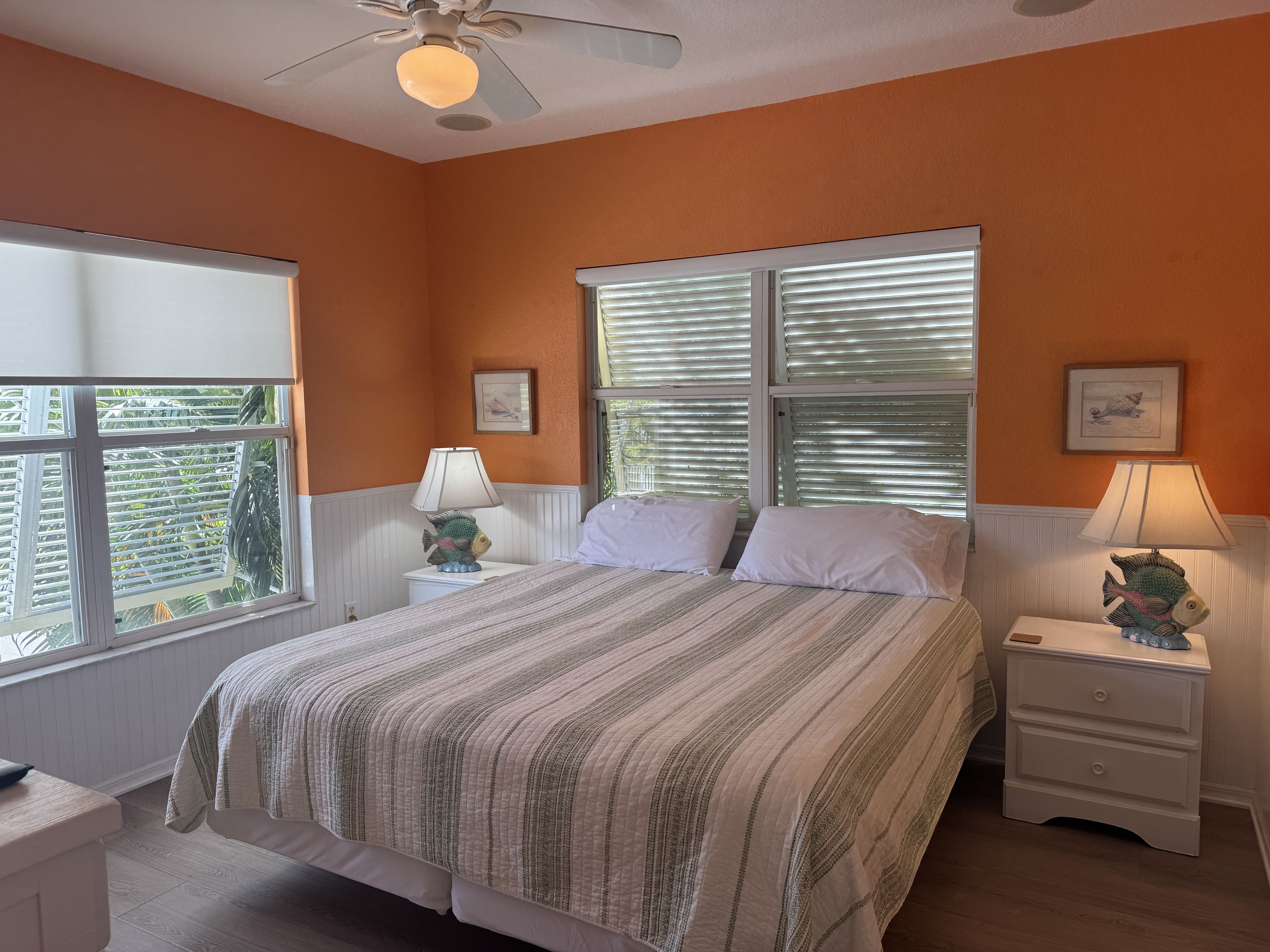 A bedroom with a bed, two bedside tables with lamps, a ceiling fan, and two windows with blinds.