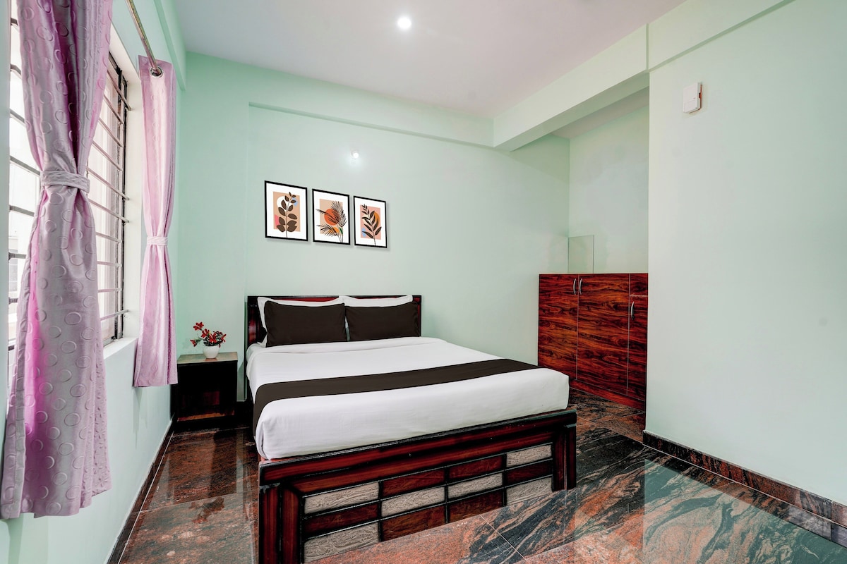 Un dormitorio con cama, mesita de noche, ventana con cortinas y una pared con tres cuadros enmarcados.