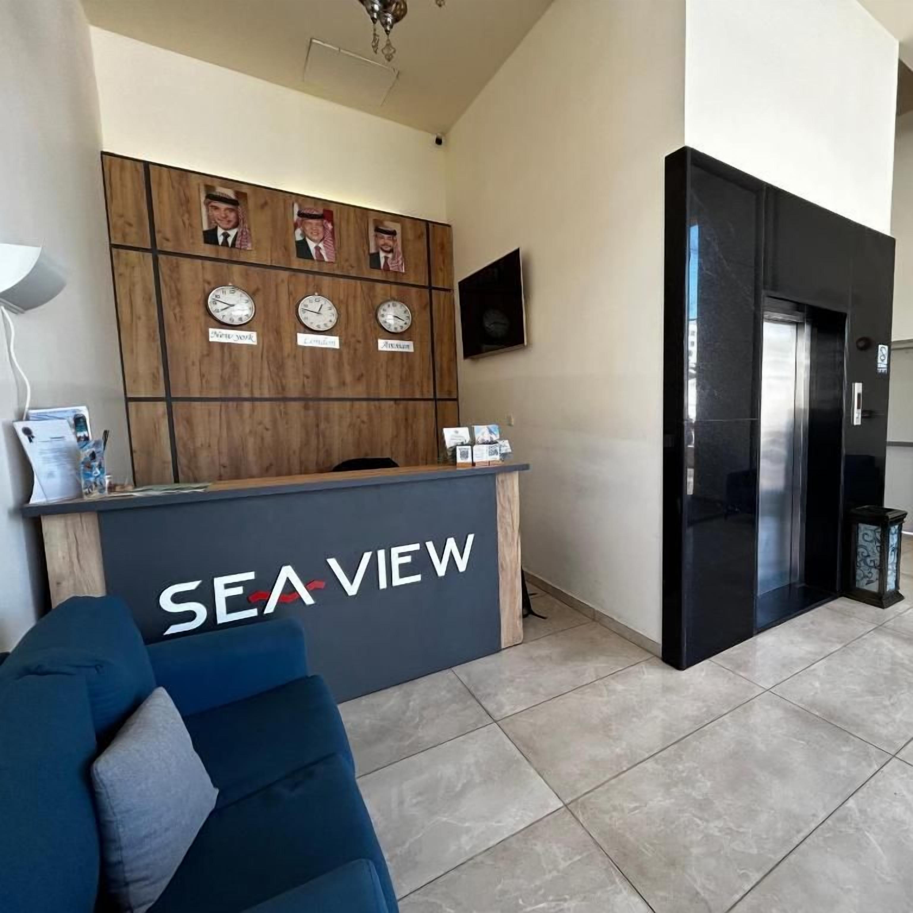A reception desk with a wooden panel design and a 'SEA VIEW' sign, a blue sofa, a wall-mounted clock, and a black elevator.