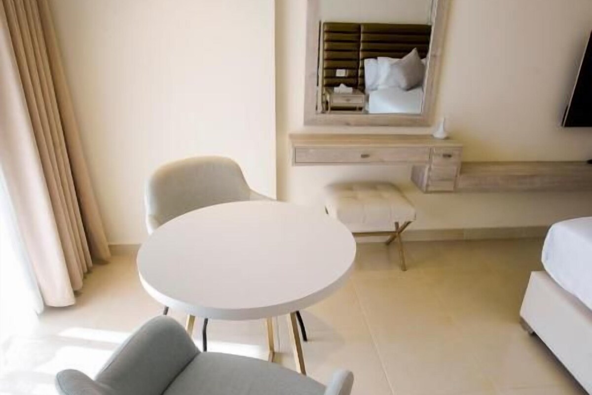 Una habitación de hotel moderna con una mesa de comedor redonda, un sillón con cojines, un sofá y un espejo grande en la pared.