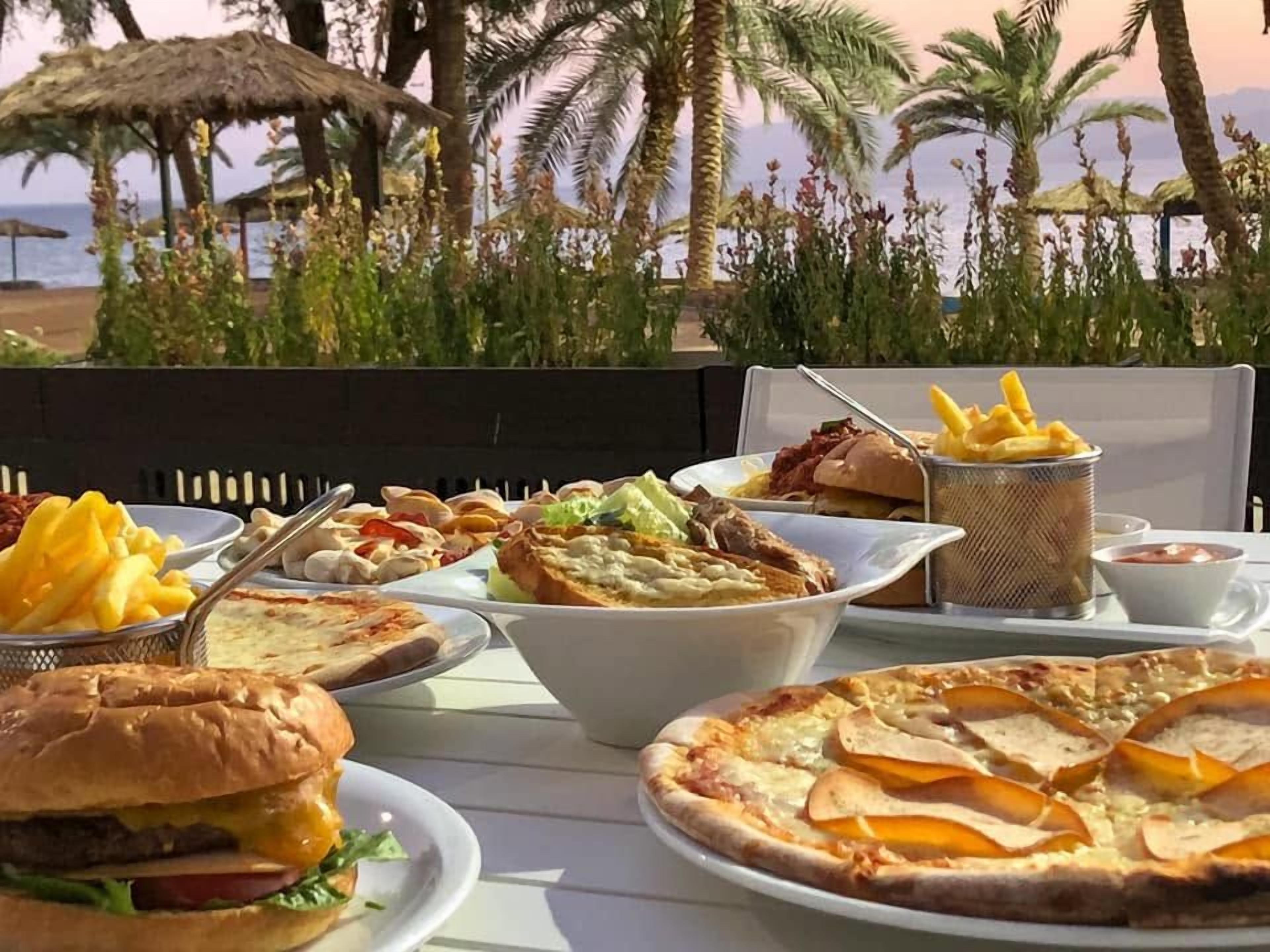 A table set with various food items including a burger, pizza, and fries, with a view of palm trees and the sea in the background.