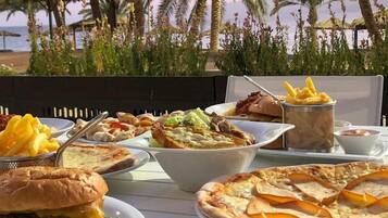 A table set with various food items including a burger, pizza, and fries, with a view of palm trees and the sea in the background.