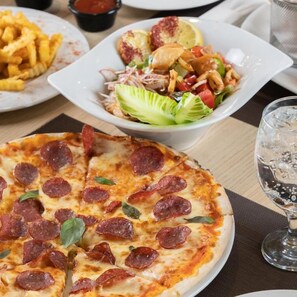 Pizza con rodajas de pepperoni, una porción de papas fritas, ensalada con vegetales y un gajo de limón, y un vaso de agua sobre una mesa.