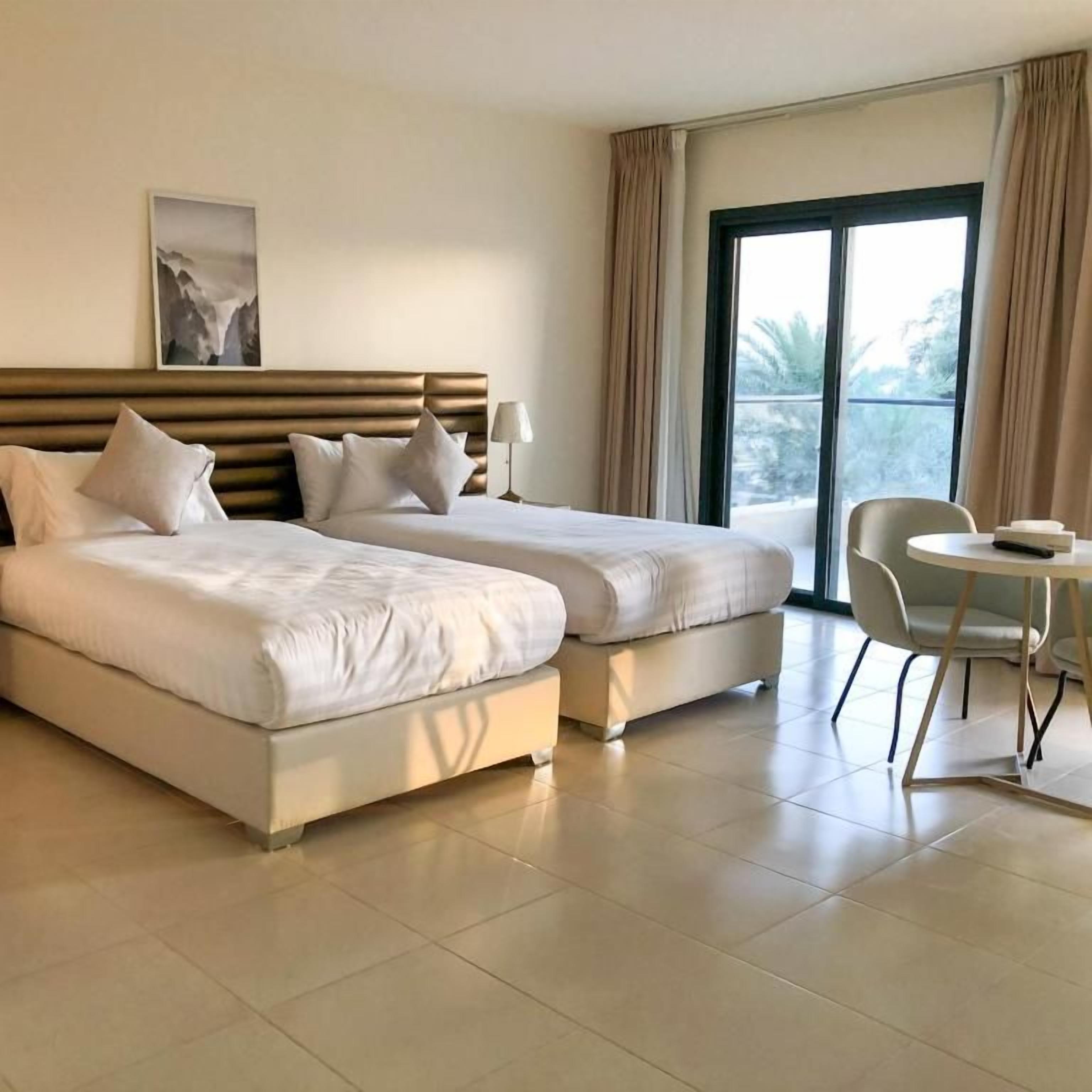 A hotel room with two beds, a chair, a table, and a large window.