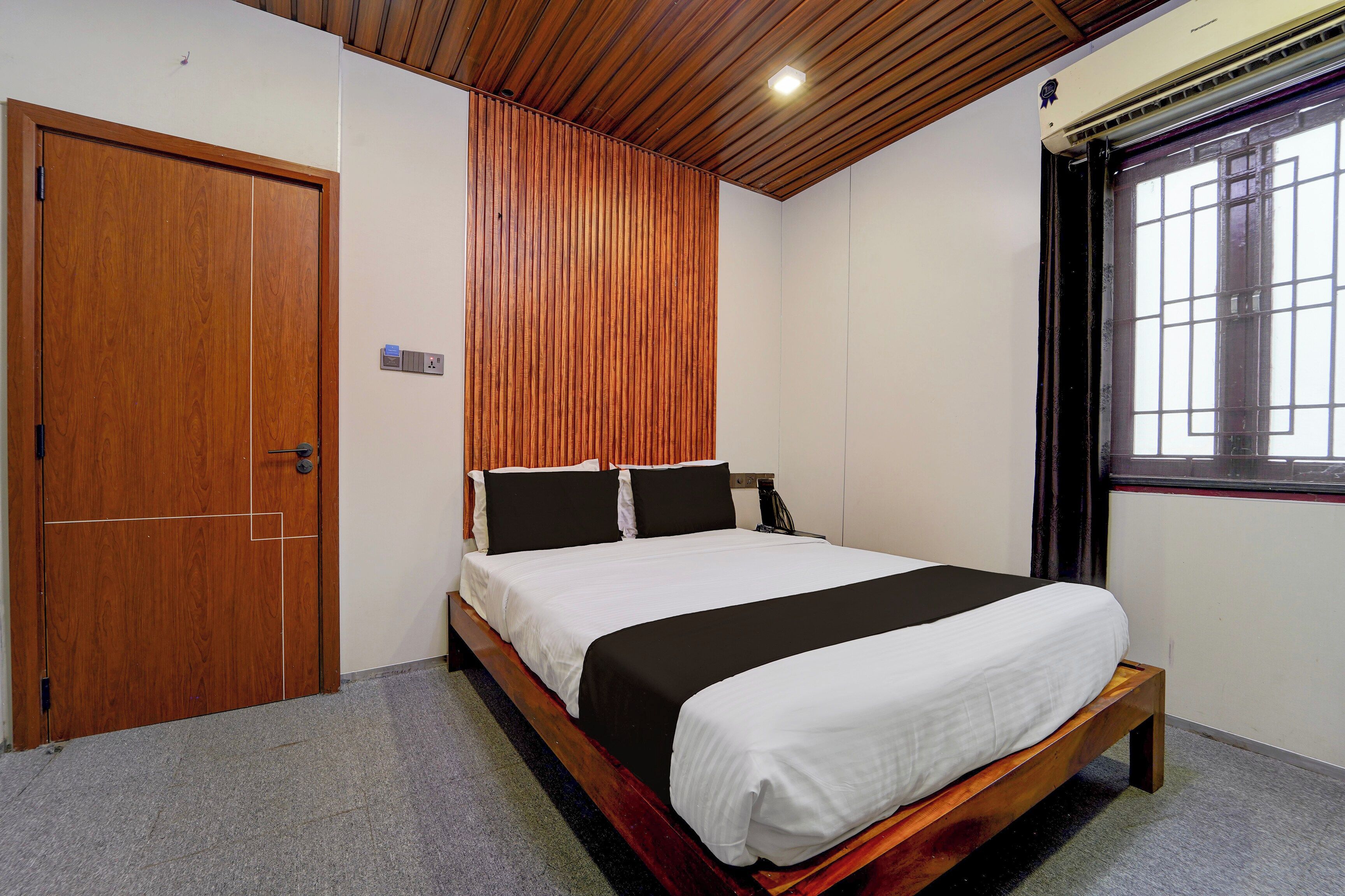 A hotel room with a wooden door, a bed with white and black bedding, a wooden headboard, and a window with a view.