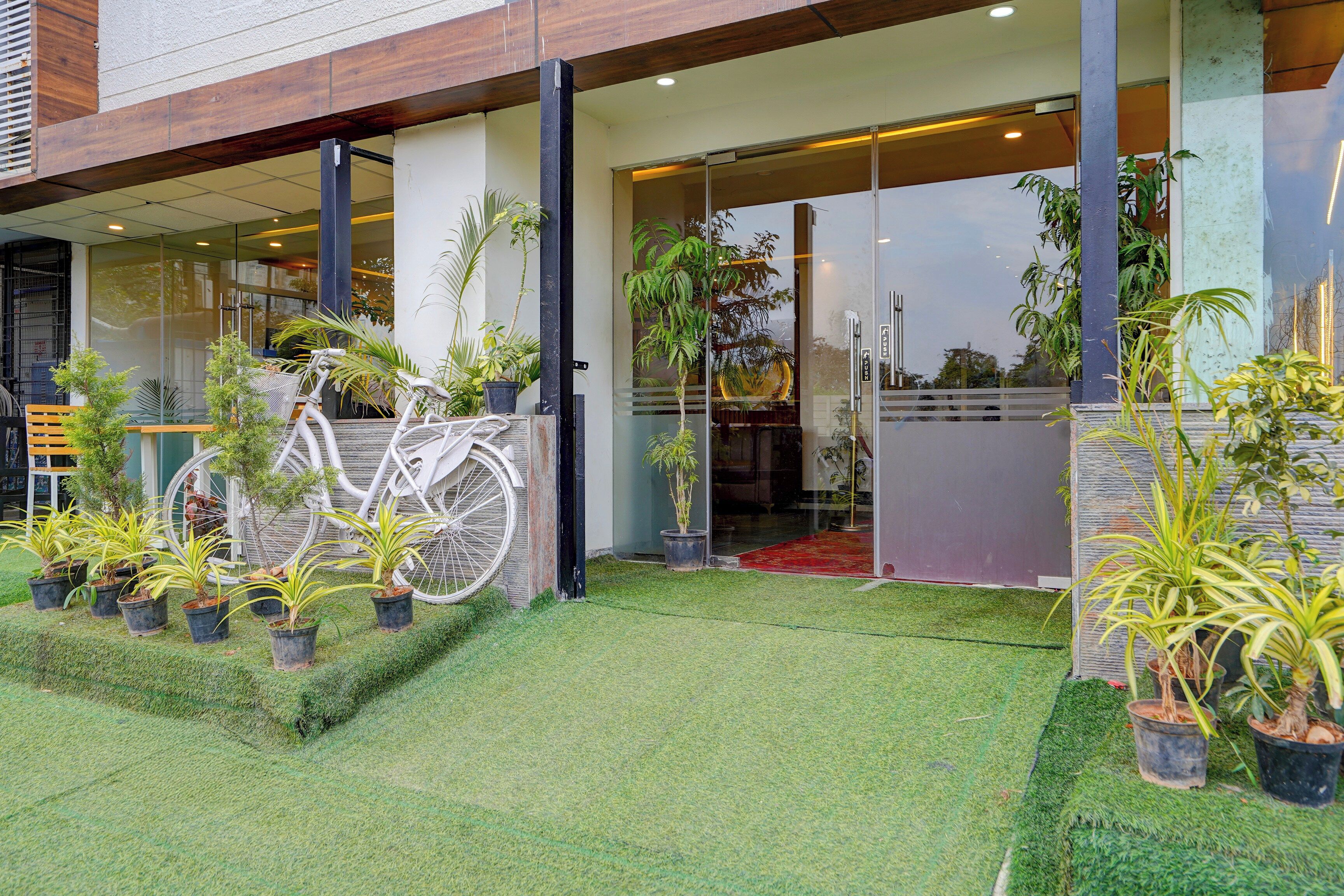 A building entrance with a bicycle parked outside, surrounded by potted plants and a grassy area.