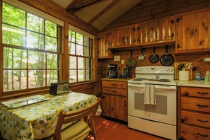 A wooden cabin kitchen with a dining table, stove, and cabinets. - New for 2026! Two Cottages on Sebago Lake w/ 160’ of Shoreline & Sunset Views (Naples)