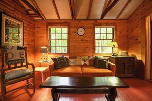 A cozy cabin interior with a wooden floor, a sofa with patterned cushions, a wooden table, a clock, and a window with a view of trees. - New for 2026! Two Cottages on Sebago Lake w/ 160’ of Shoreline & Sunset Views (Naples)