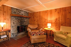 A cozy living room with a stone fireplace, wooden walls, a leather armchair, a small ottoman with a quilt, and two table lamps. - New for 2026! Two Cottages on Sebago Lake w/ 160’ of Shoreline & Sunset Views (Naples)