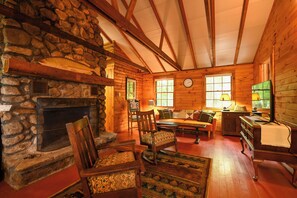 A log cabin interior with a stone fireplace, wooden furniture, and a large window. - New for 2026! Two Cottages on Sebago Lake w/ 160’ of Shoreline & Sunset Views (Naples)