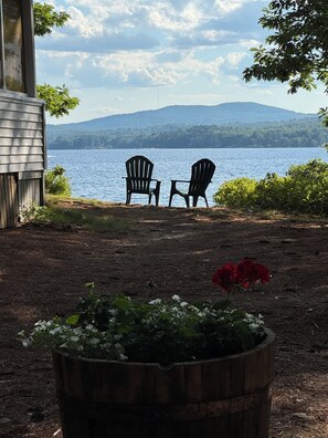 Vista sul lago con due poltrone Adirondack rivolte verso l'acqua, un pontile in legno e un'aiuola in primo piano.