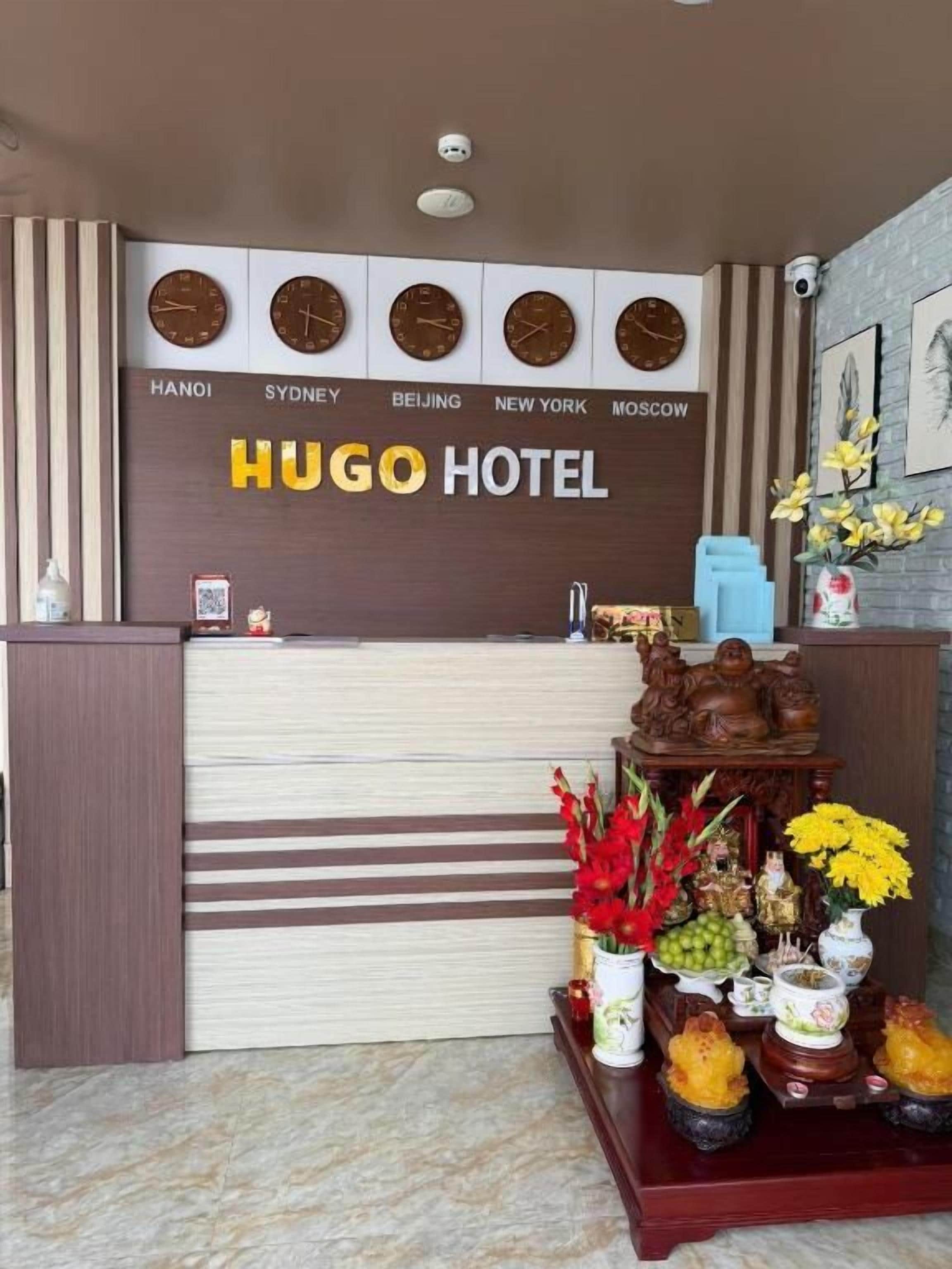 A reception desk with clocks displaying times from various cities, a Hugo Hotel sign, and a decorative display with flowers and fruits.