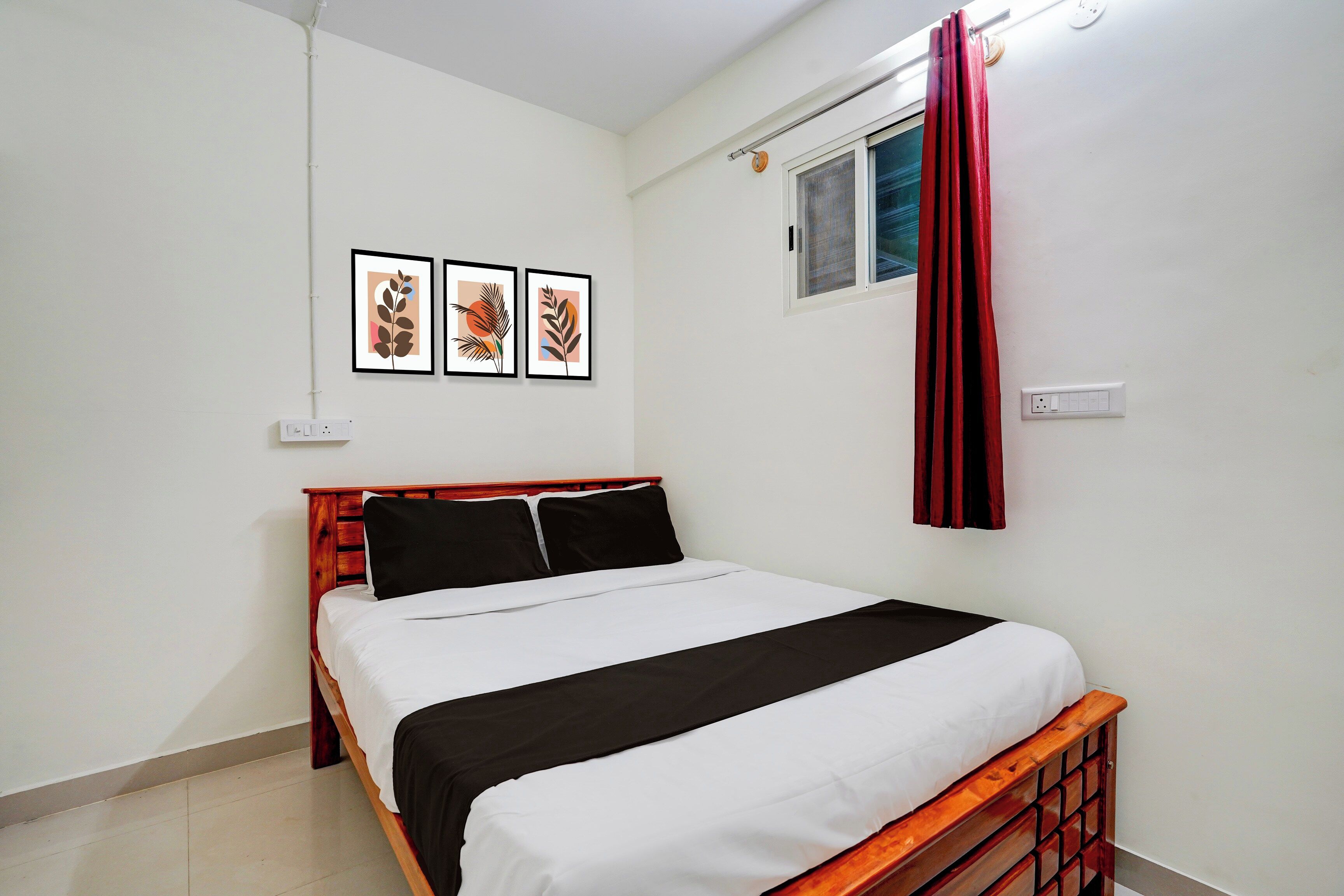 A bedroom with a wooden bed, white bedding, a window with red curtains, and three framed artworks on the wall.