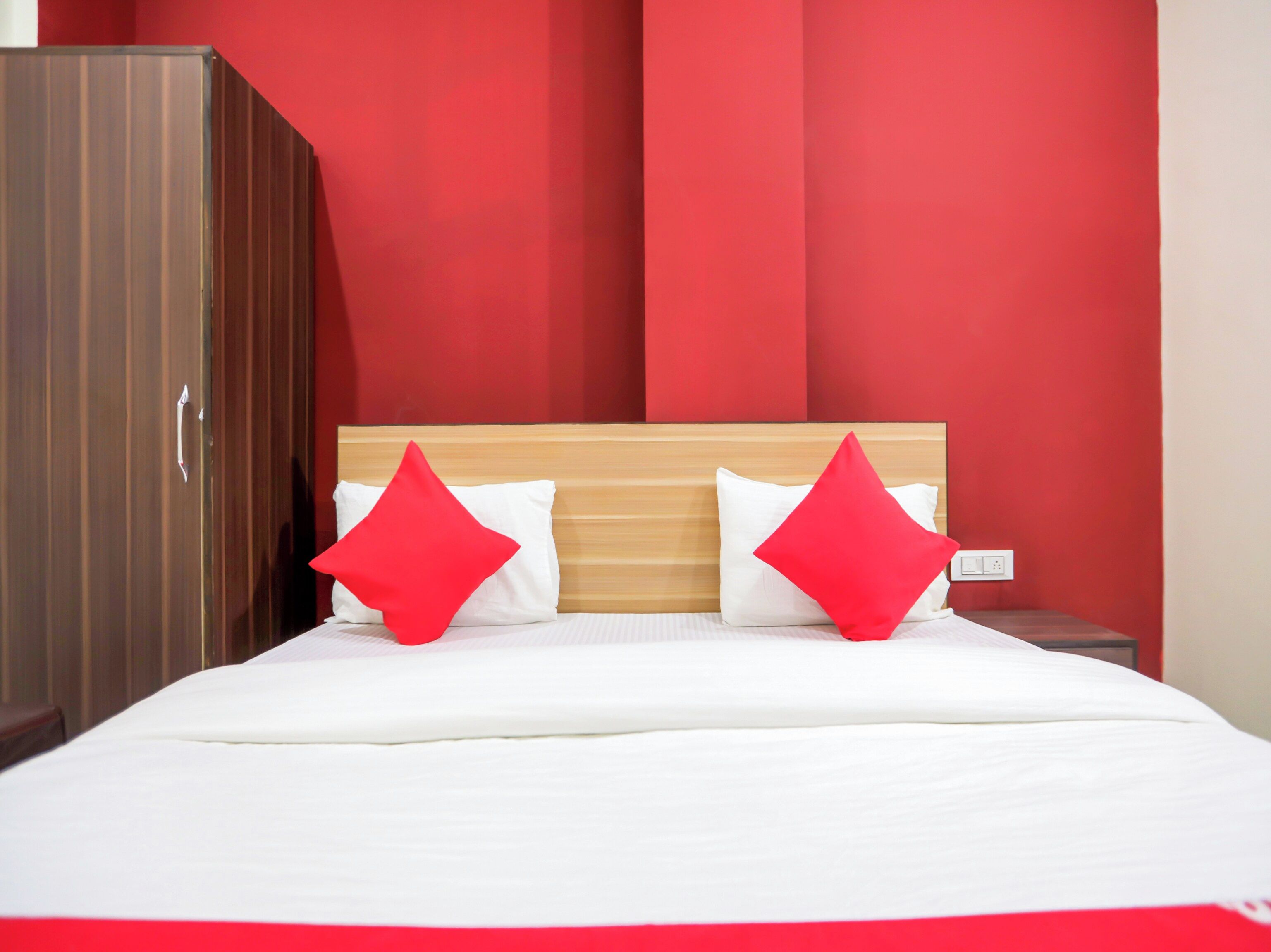 Una cama con ropa de cama blanca y dos almohadas rojas, cabecera de madera y un armario con acabado marrón.