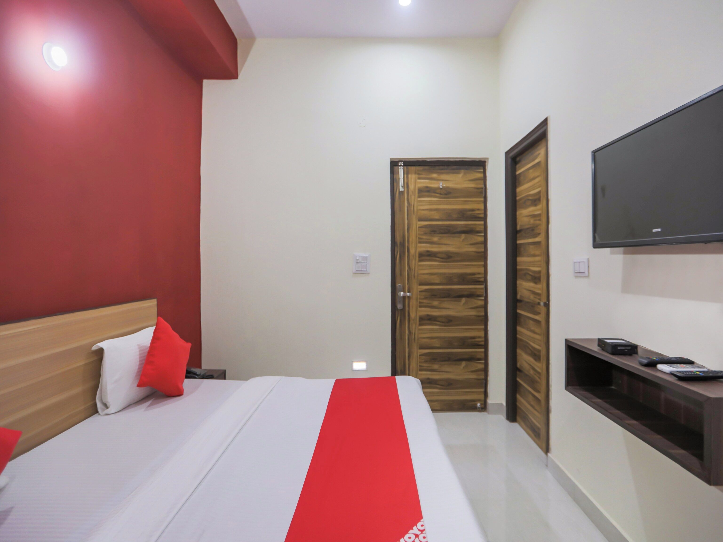 A hotel room with a bed, a TV mounted on the wall, a wooden door, and a small table with a remote control.