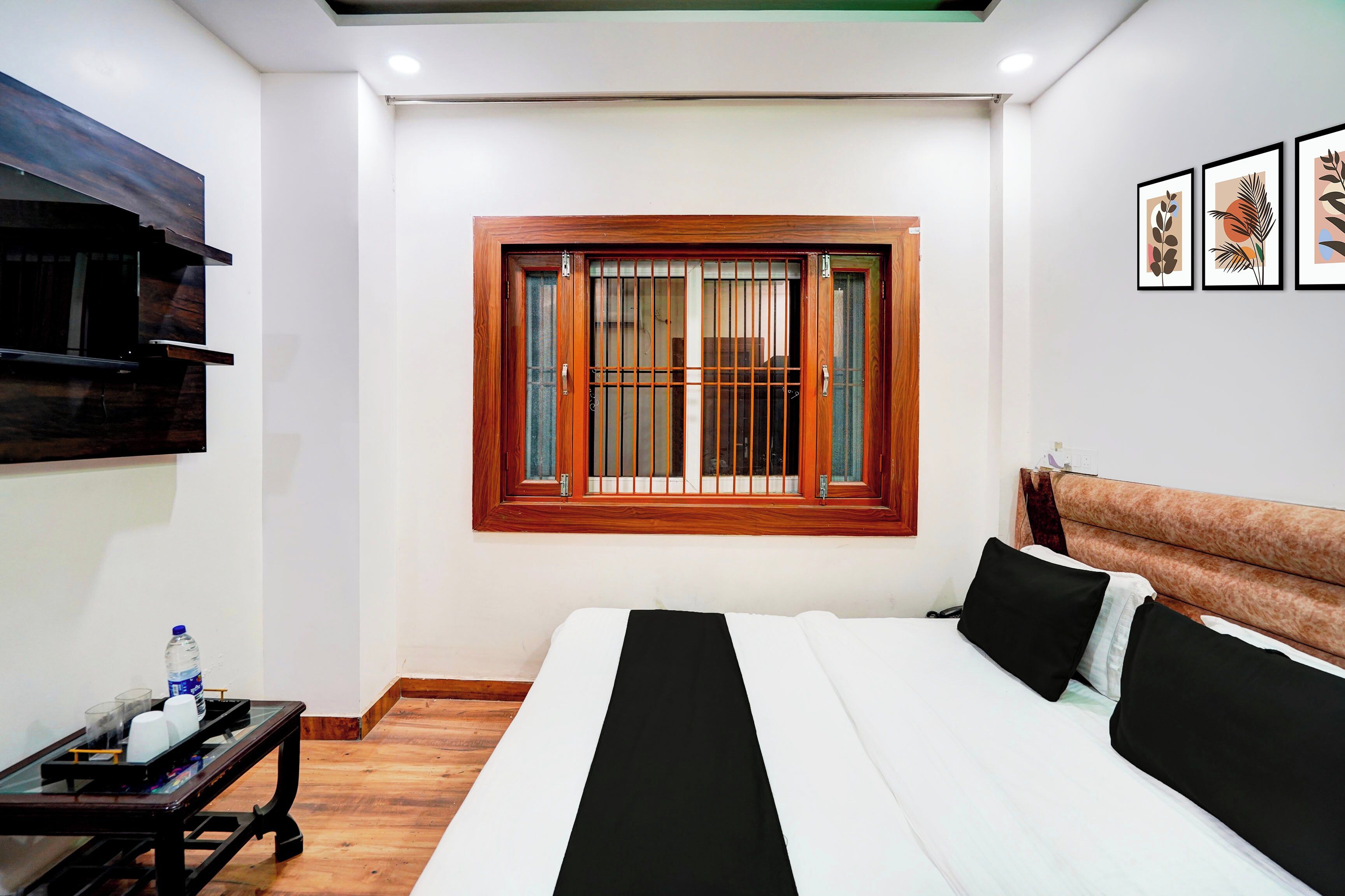 A hotel room with a bed, a small table, a window with bars, and framed artwork on the wall.