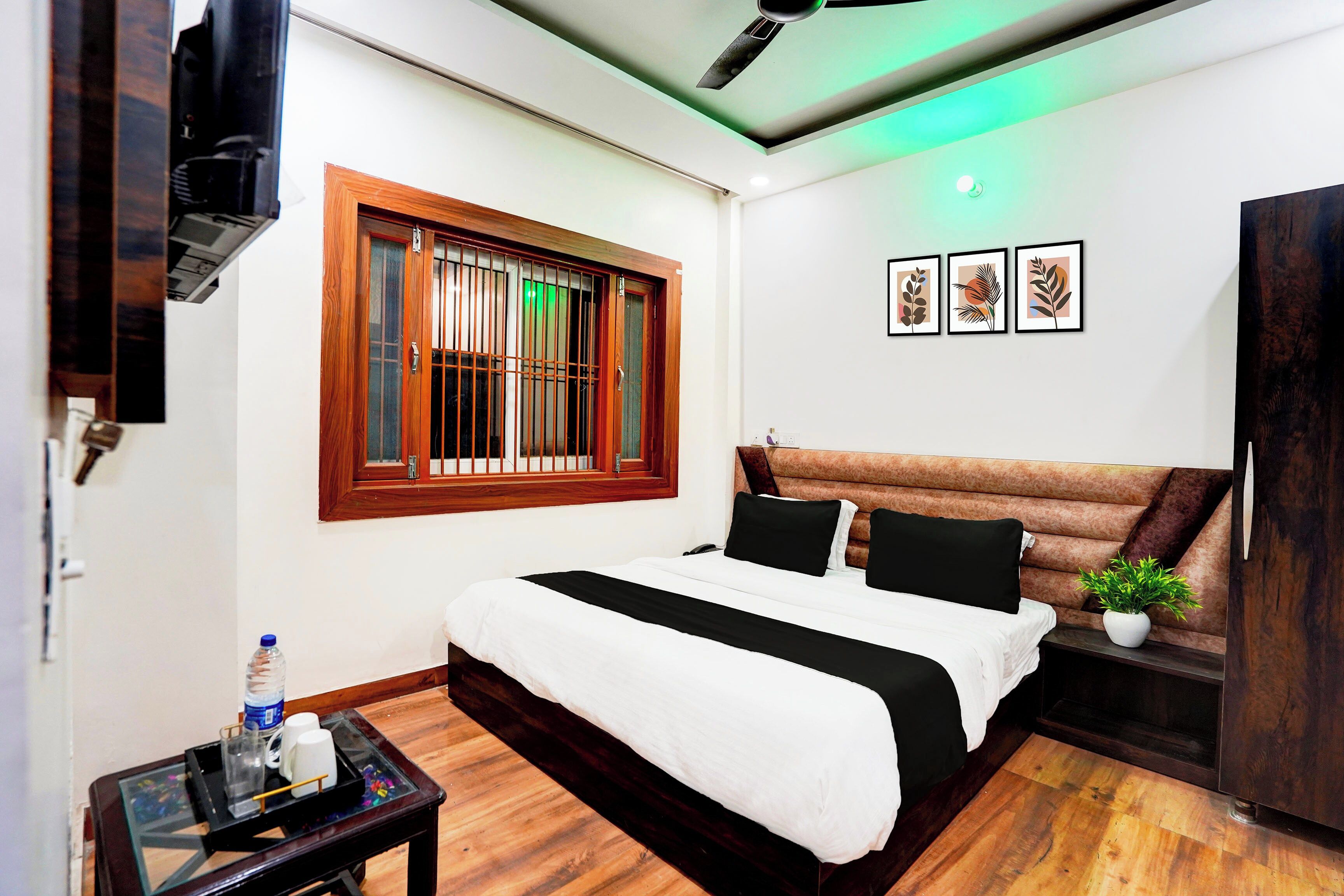 A hotel room with a bed, a small table with a bottle and glasses, a window with bars, and a ceiling fan.