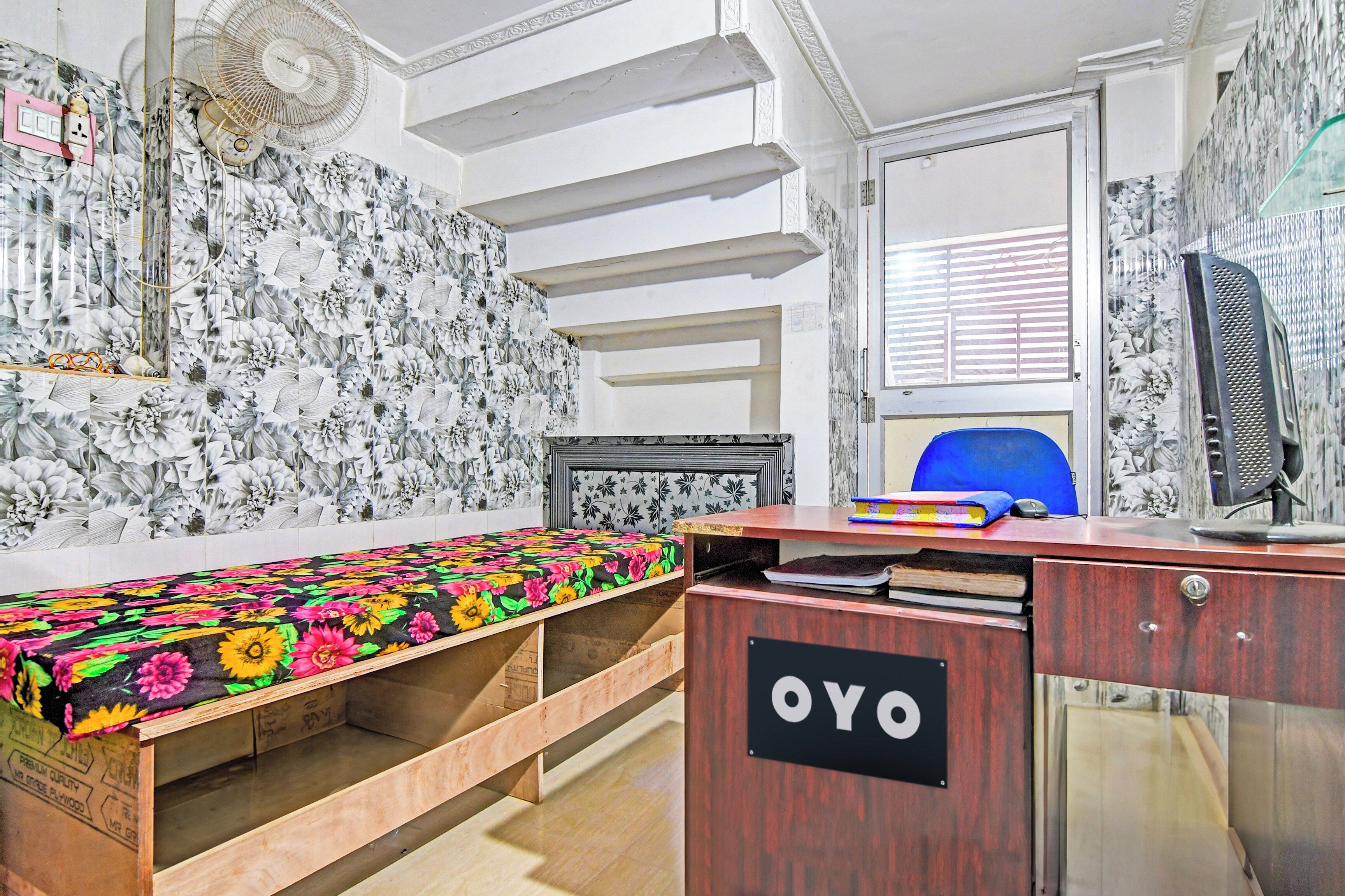A room with a bed, a desk with a computer, a chair, and a wall with floral wallpaper.