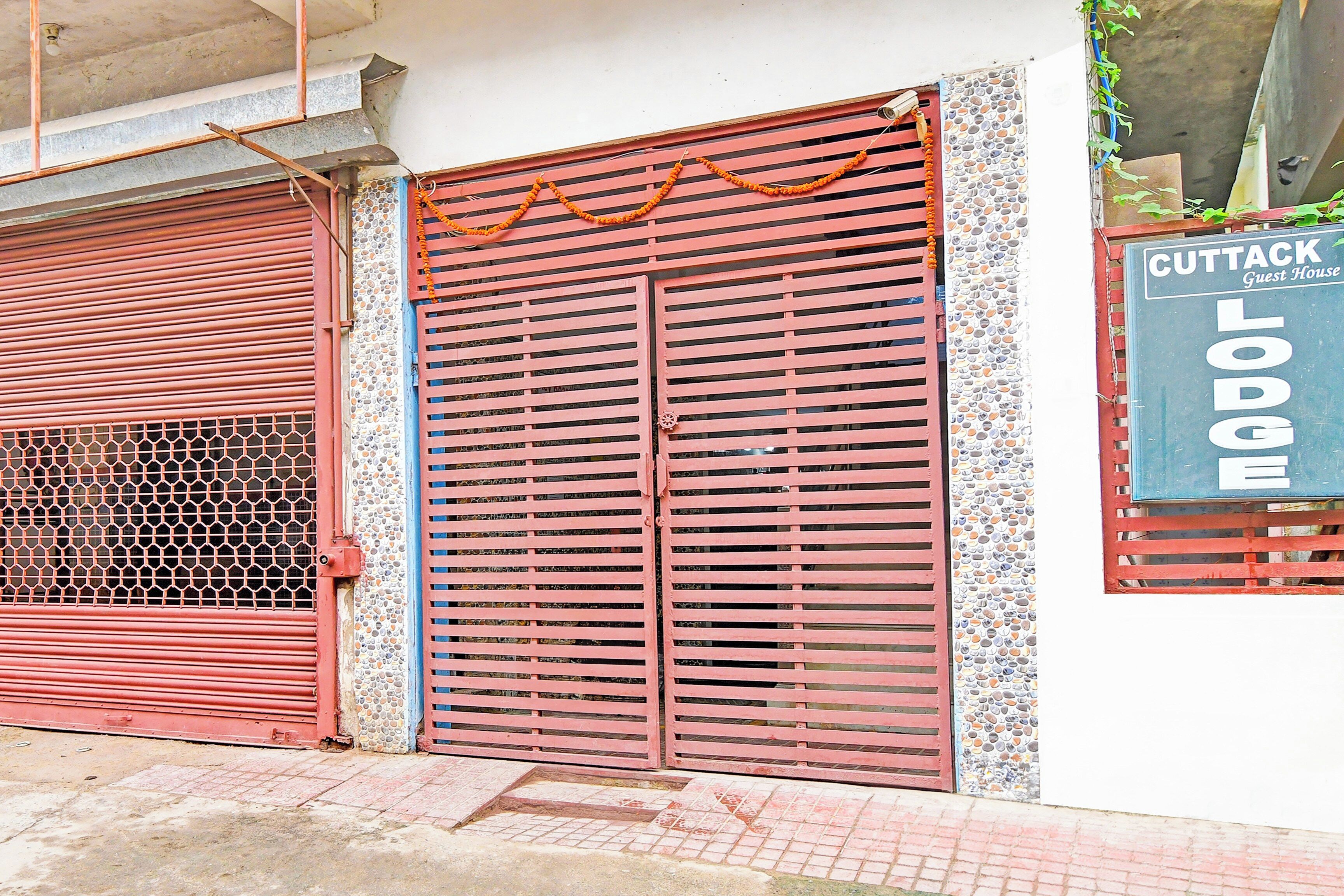 A building with two red metal shutters, a signboard for 'Cuttack Guest House Lodge', and a decorative garland.