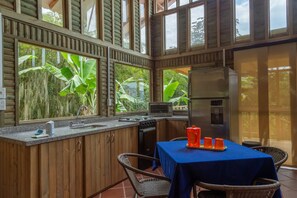 A kitchen with a sink, stove, and refrigerator, a dining table with chairs, and large windows with a view of greenery. - LA CABAÑA EN The GUADUAL (San Antonio Del Tequendama)
