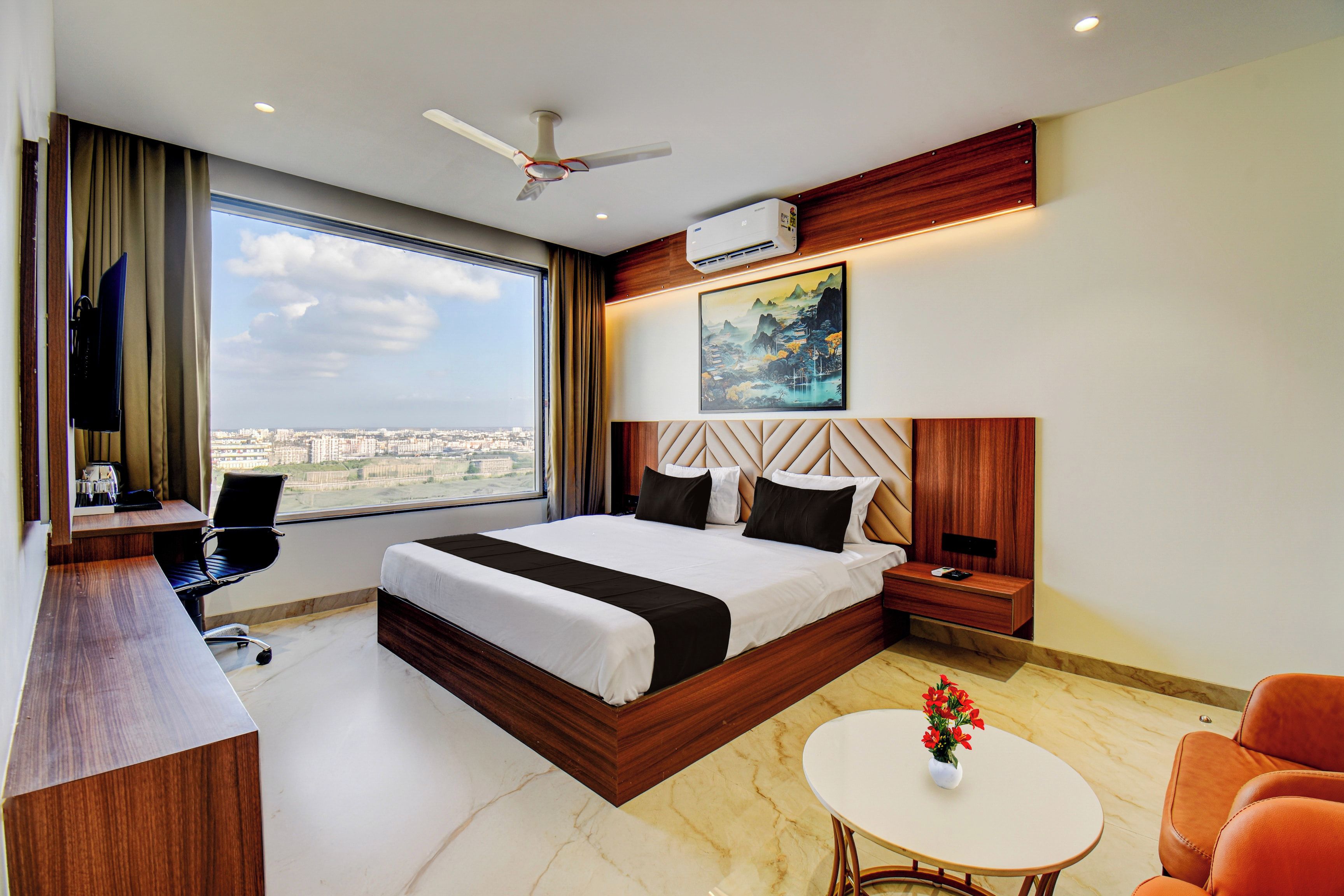 Habitación de hotel con una cama grande, un escritorio, una silla, una pequeña mesa con flores y vistas a la ciudad.