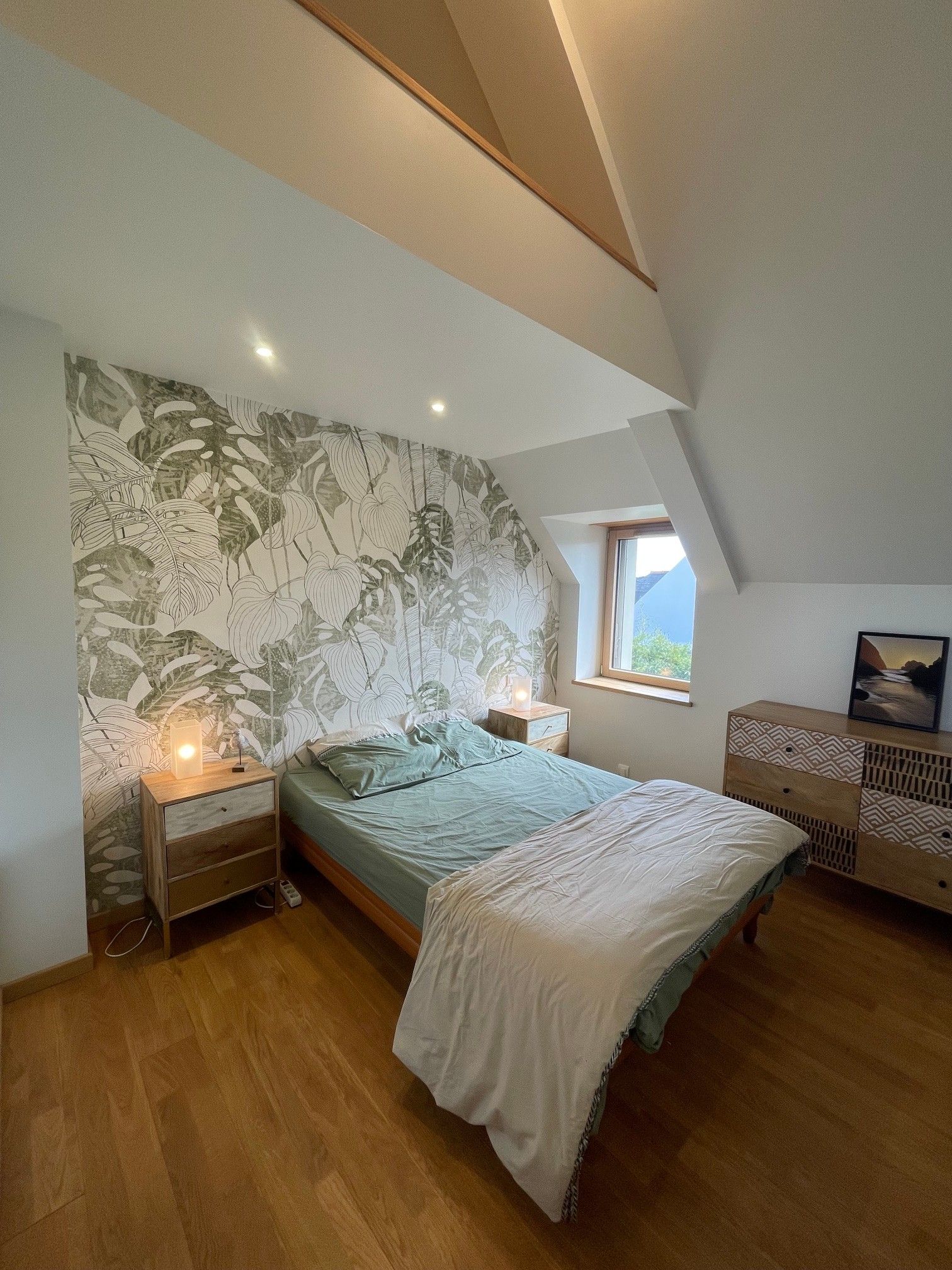 A bedroom with a sloped ceiling, a bed with a patterned bedspread, a bedside table with a lamp, and a window with a view.