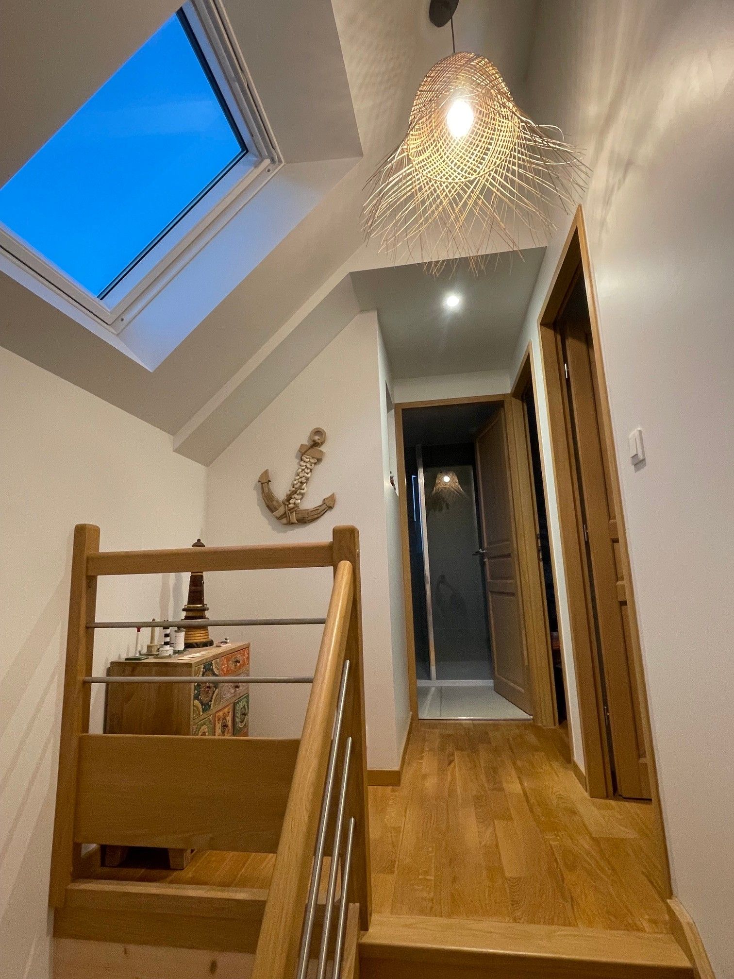 Un escalier avec une rampe en bois mène à un palier où se trouve une petite table et des objets décoratifs. Une lucarne est visible au-dessus du palier, et un luminaire tressé est suspendu au plafond.