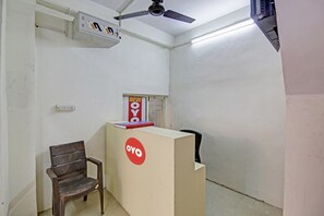 A reception desk with a sign, a wooden chair, and a ceiling fan in a room with white walls. - Hotel O ROYAL 18 (Noida)