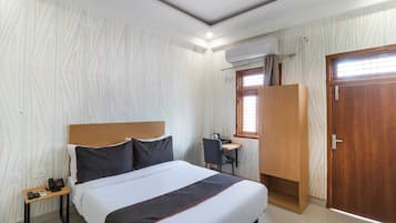A hotel room with a bed, bedside table, desk, chair, wardrobe, and an air conditioning unit.